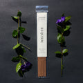 Requiem Incense Company Cardamom & Tobacco incense sticks with green leaves and purple flowers on a dark background